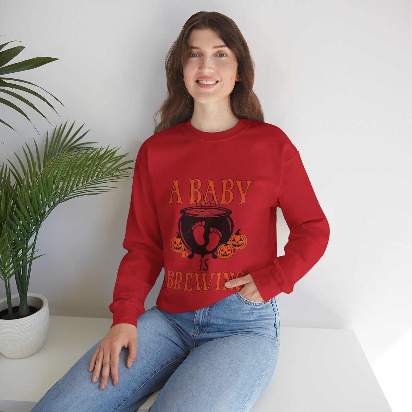 Brewing Sweetshirt maternity Halloween pregnancy announcement sweatshirt in red worn by a woman sitting near plants.