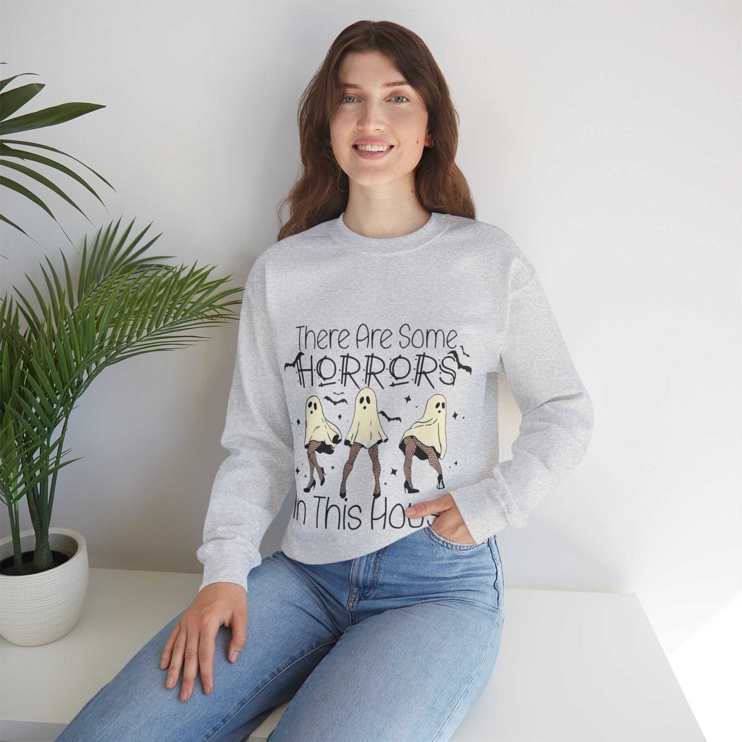 Funny Halloween sweatshirt with "There Are Some Horrors in This House" design, worn by a smiling person sitting on a white surface, surrounded by greenery.