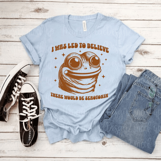 Funny Frog Tee with "I Was Led to Believe There Would Be Serotonin" design on light blue shirt, paired with jeans and sneakers.